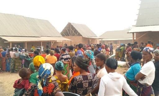 A group of people wearing colorful clothing gather outdoors in front of several buildings with corrugated metal roofs.